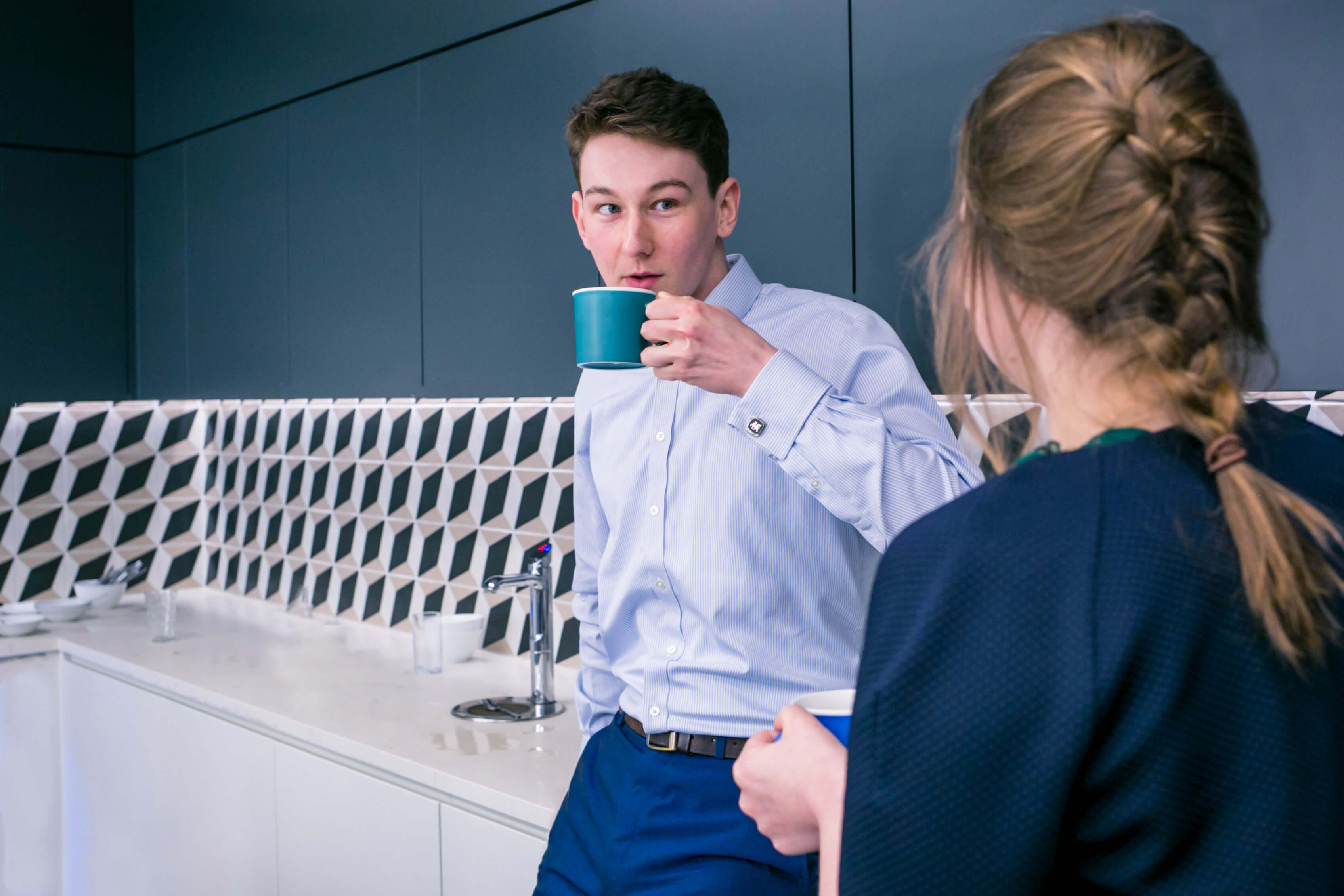 Two colleagues chatting and drinking a beverage in an office kitchen. The male colleague is in focus but you can only see the female colleague's back