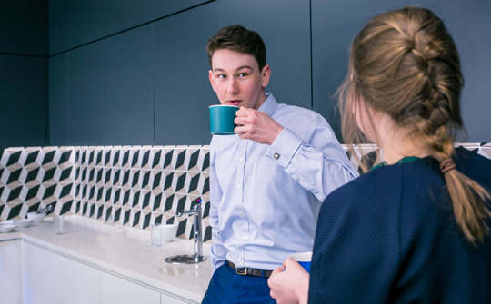 Two colleagues chatting and drinking a beverage in an office kitchen. The male colleague is in focus but you can only see the female colleague's back