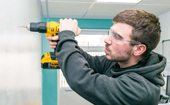 A man drilling a hole in a white wall. He's wearing a grey jumper and protective glasses