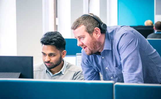 Two colleagues in front of a desk. They're focused on something on the computer screen in front of them while they're chatting.