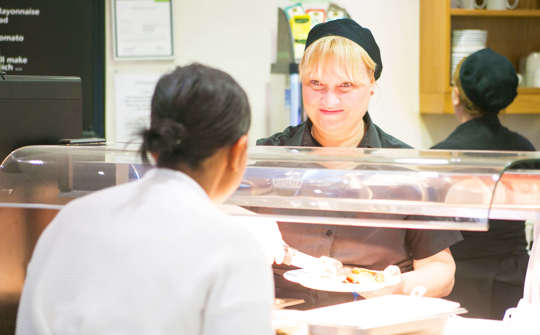 Woman serving food in a canteen. She's smiling at the person she's serving.