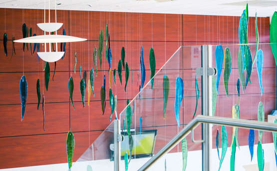 Artwork with a school of fish hanging from a ceiling. The wall behind it has a warm auburn colour. The fish are a mixture of colours - either blue or green. The depth of colour changes depending on the light.