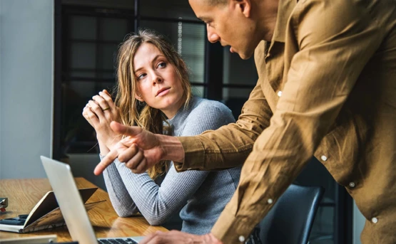 Two colleagues at a table. The male colleague is talking and pointing to something on the screen. The female colleague is listening