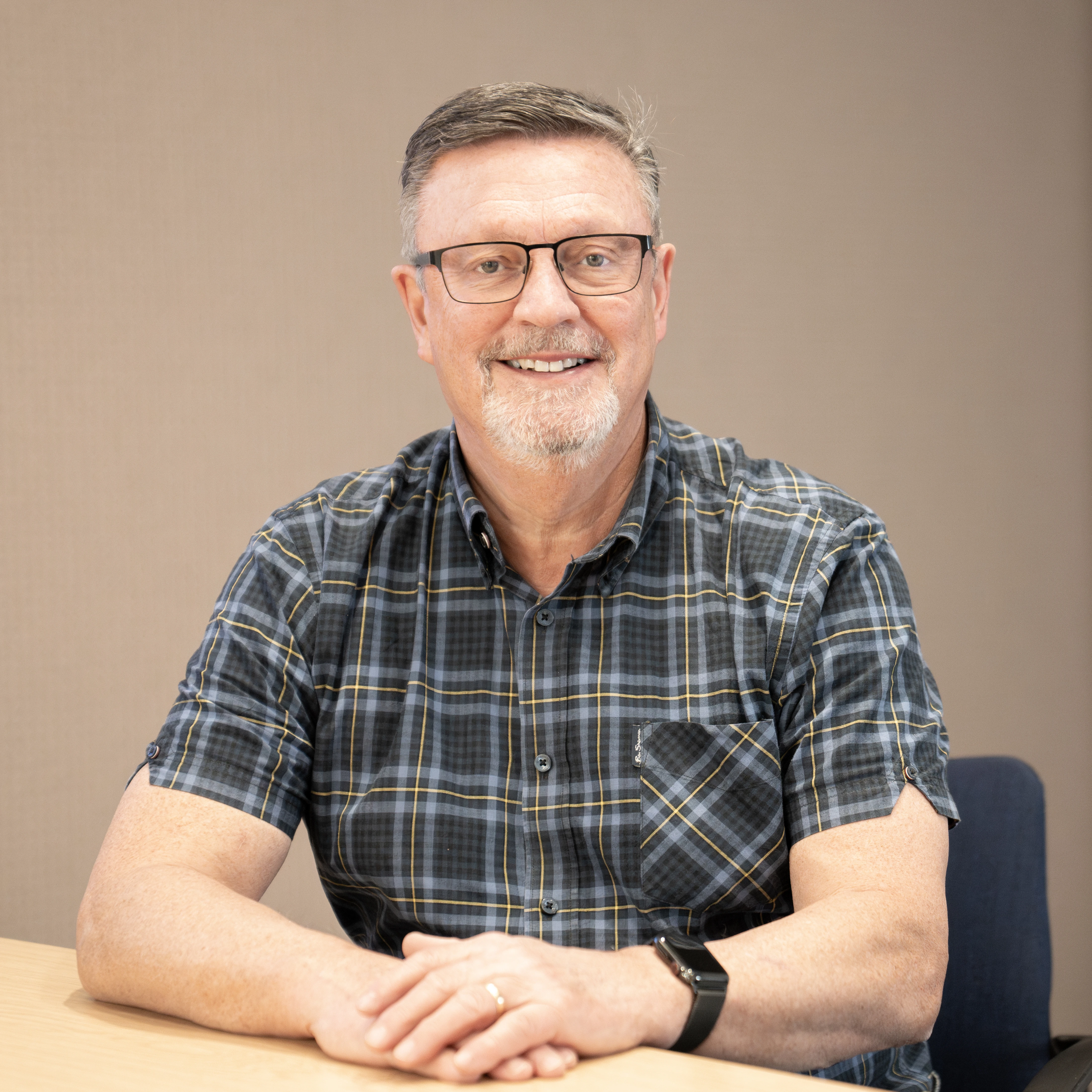 A smiling middle-aged man with glasses and a short beard sits at a table. He is wearing a short-sleeved, plaid button-up shirt and is positioned against a plain beige background.