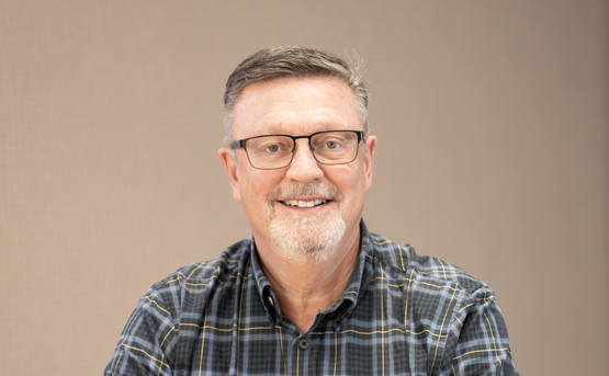 A smiling middle-aged man with glasses and a short beard sits at a table. He is wearing a short-sleeved, plaid button-up shirt and is positioned against a plain beige background.