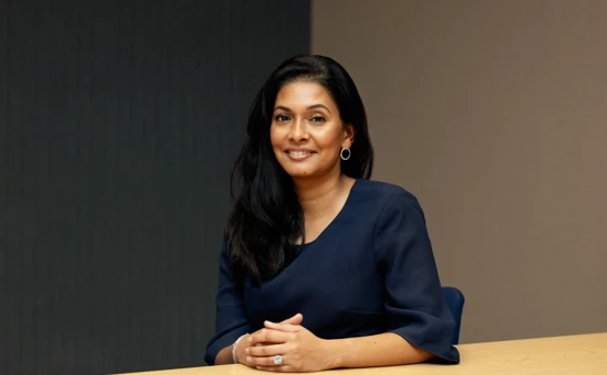 A woman with long dark hair, wearing a dark blue dress, sits with her hands clasped at a wooden desk.