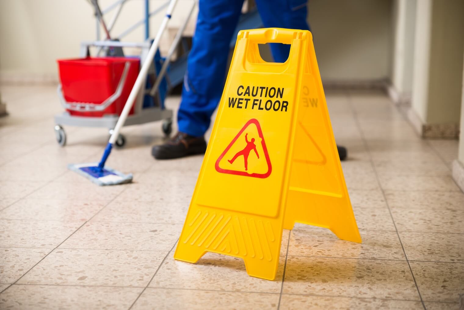 A bright yellow wet floor sign standing as a worker mops the floor behind 