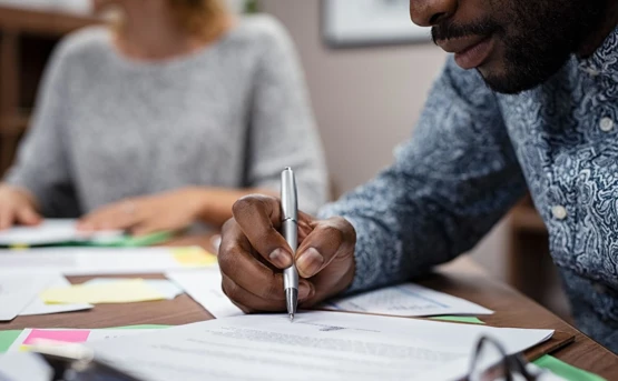 a man in a floral shirt signs some paperwork with a silver pen in front a woman