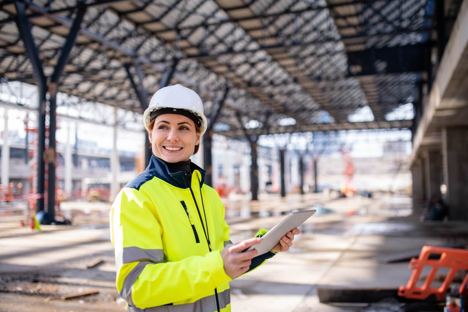 a woman wearing  a high-vis jacket and white safety helmet holds a tablet and is smiling on a construction site