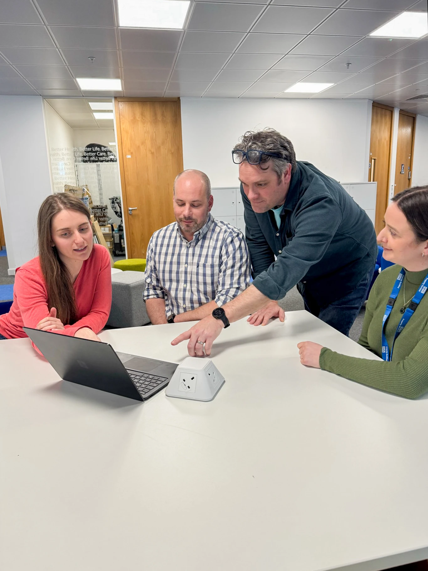 Four people sit around a table looking at a laptop. One man stands and points at the screen while the others watch attentively. They are in a modern office space with wooden doors and bright lighting.