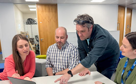 Four people sit around a table looking at a laptop. One man stands and points at the screen while the others watch attentively. They are in a modern office space with wooden doors and bright lighting.