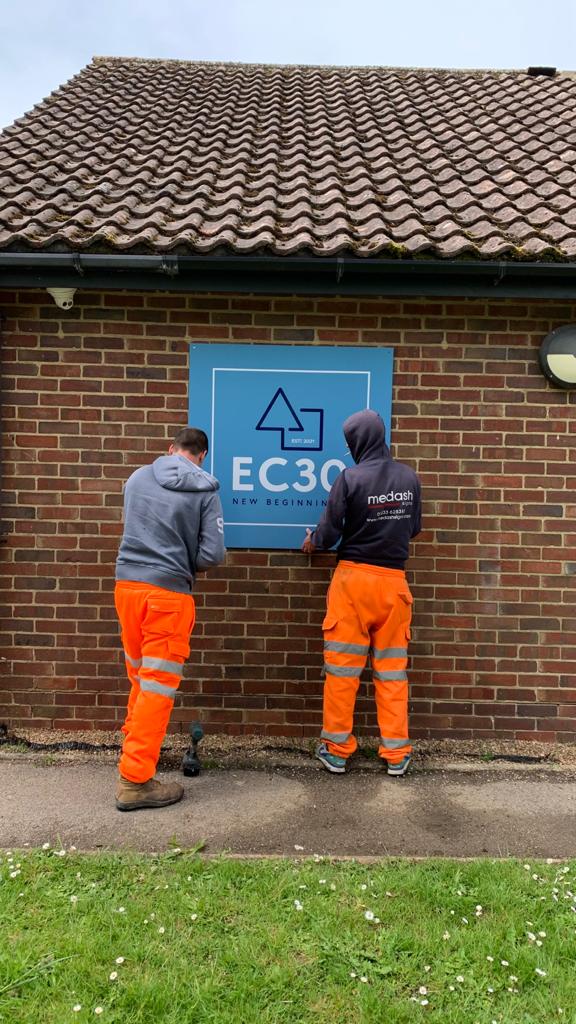 Two men in orange high-vis trousers putting up EC30 sign on side of brick wall