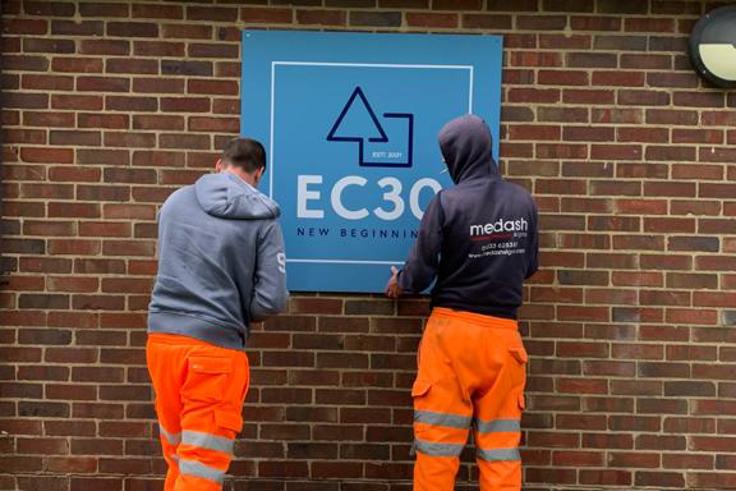 Two men in orange high-vis trousers putting up EC30 sign on side of brick wall