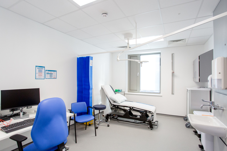 A GP exam room in Kier Devizes Health Centre. The room has a desk with a computer and a blue office chair. A bed is behind a blue privacy curtain and a sink. 