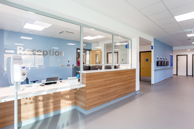 The reception desk at Kier Devizes Health Centre. 'Reception' is written in large silver letters on a blue wall. There's a glass screen in front of the reception desk which has four monitors and keyboards. 