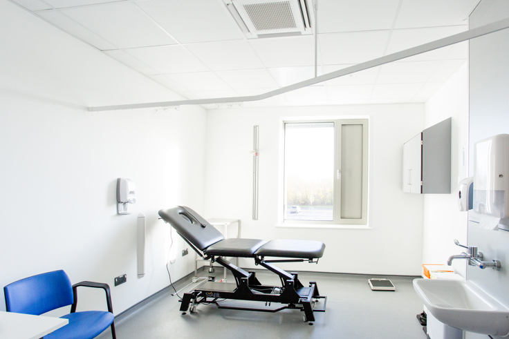 An exam room in Devizes Health Centre. The room has a black patient bed, seating and a sink. The room is decorated white and blue. 
