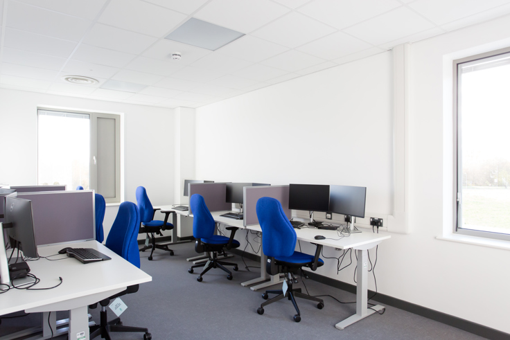 An office working area in Devizes Health Centre. There are deskspaces partitioned by grey screens. Each desk space has two monitors, a keyboard, a white desk and a blue chair. 