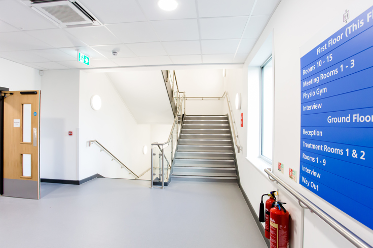 A hallway in Devizes Health Centre. There are two flights of stairs to the next floor and a blue and white sign.
