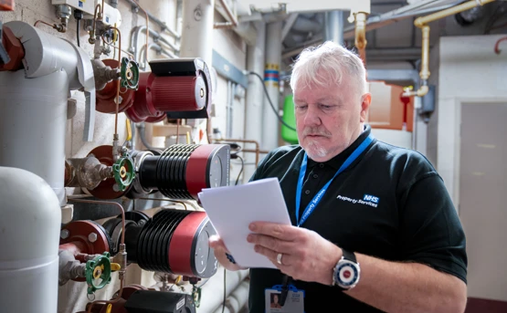 An older man in an NHS uniform inspects machinery and reads a clipboard in an industrial room with pipes and gauges.