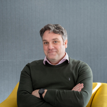 A man with short, grayish hair sits in a yellow chair, arms crossed, wearing a green sweater over a collared shirt, against a gray textured background.