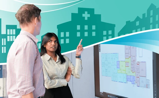 A woman points at a wall display with a building layout while talking to a man. Behind them is a graphic of hospital buildings with a medical cross symbol.
