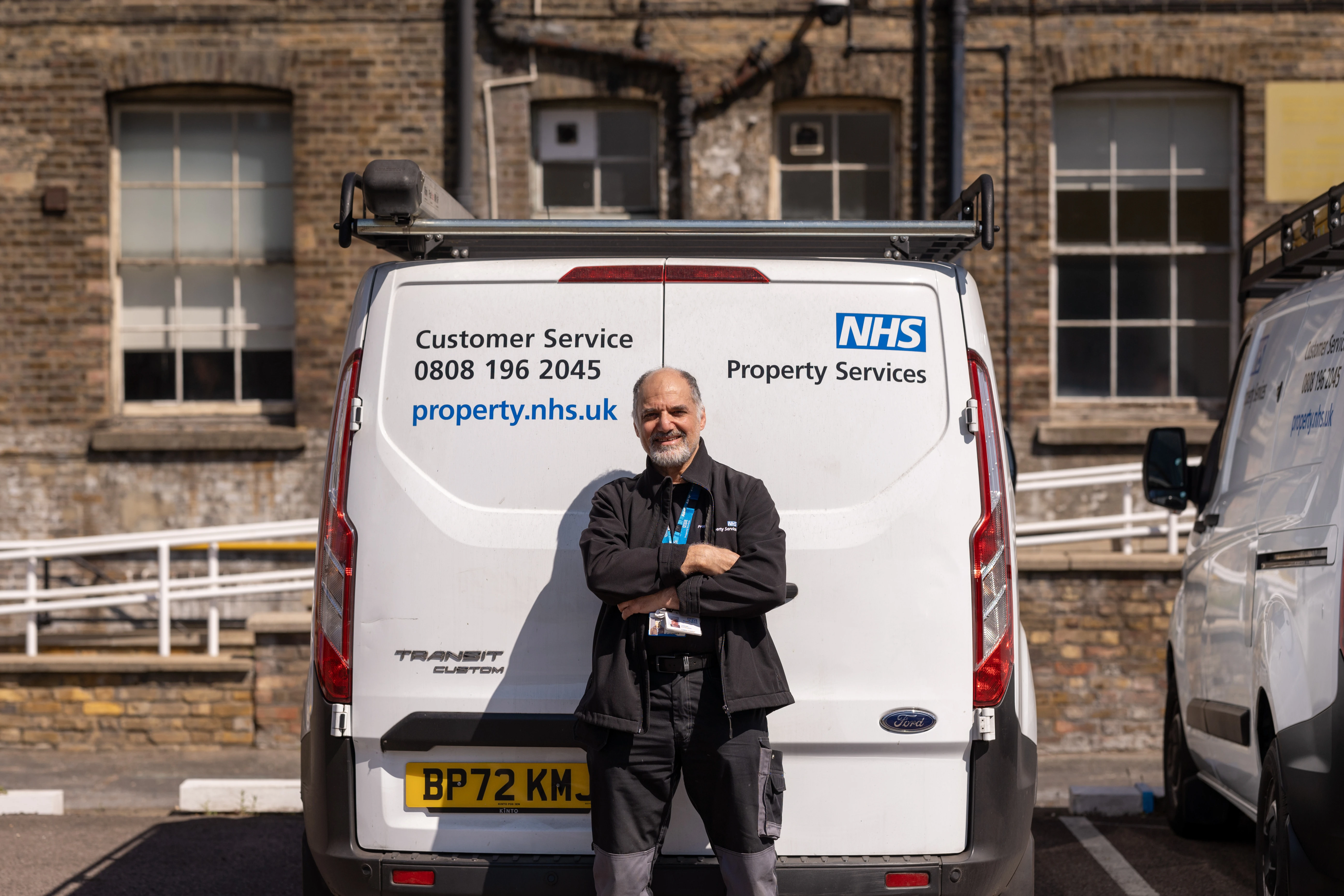 A man stands smiling with arms crossed in front of an NHS Property Services van in a sunny parking lot, with a brick building in the background.