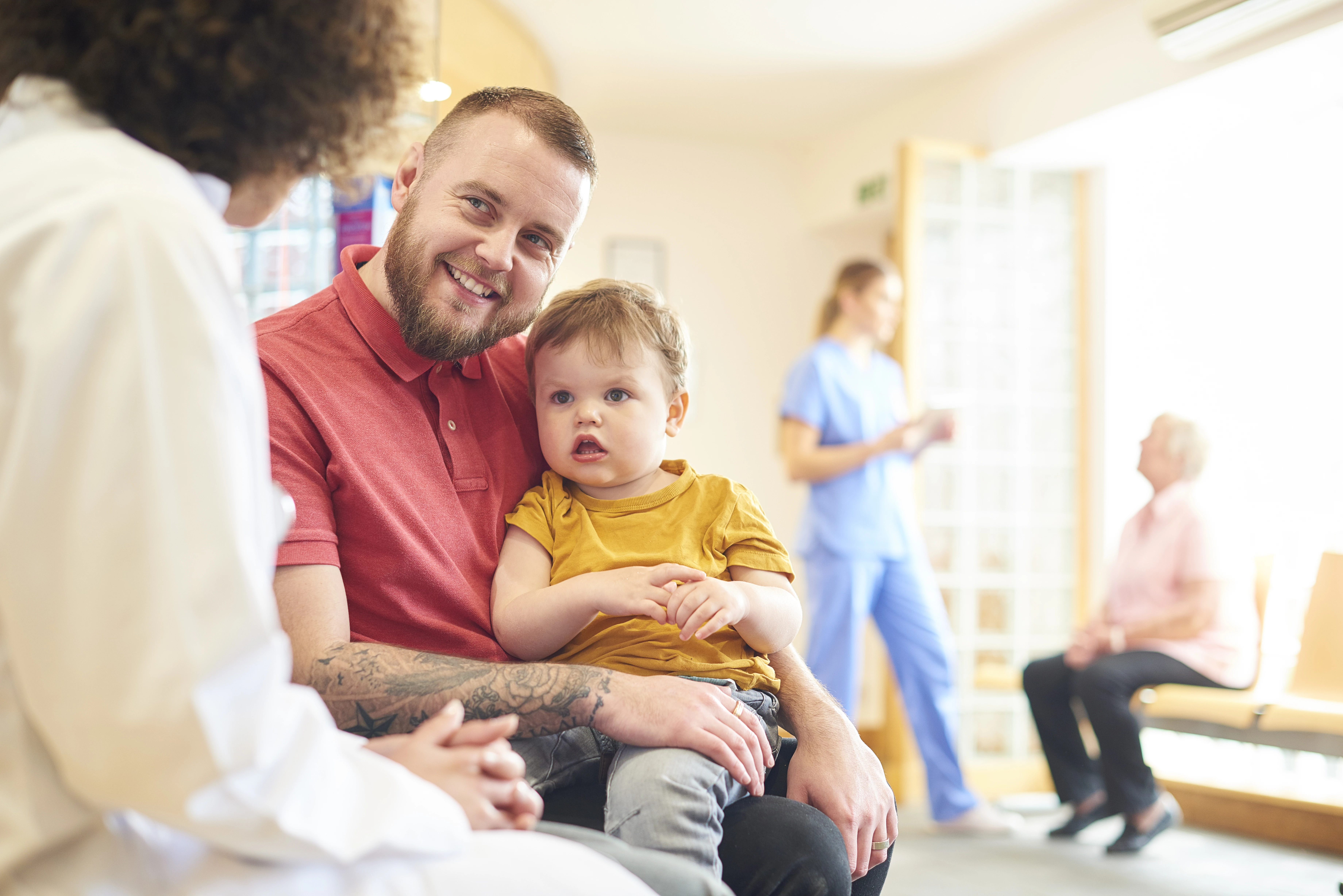 A man in a red shirt holds a young child in his lap while talking to a healthcare worker. Two more people, one in scrubs and one elderly, are in the background of a bright waiting room.