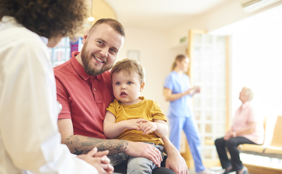 A man in a red shirt holds a young child in his lap while talking to a healthcare worker. Two more people, one in scrubs and one elderly, are in the background of a bright waiting room.