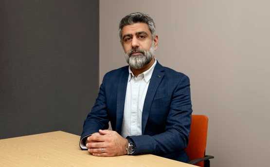 A middle-aged man with a graying beard, wearing a navy blue blazer and white dress shirt, sits at a wooden table with his hands clasped.