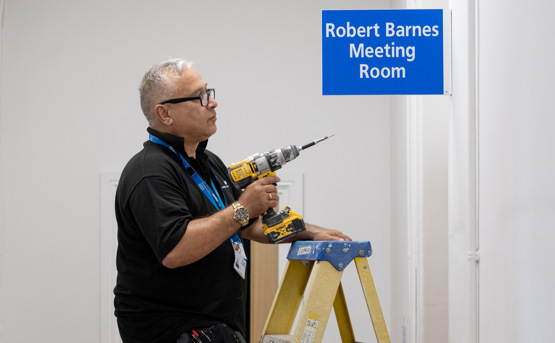 A man stands on a ladder using a power drill near a blue sign that reads Robert Barnes Meeting Room in a white-walled room.
