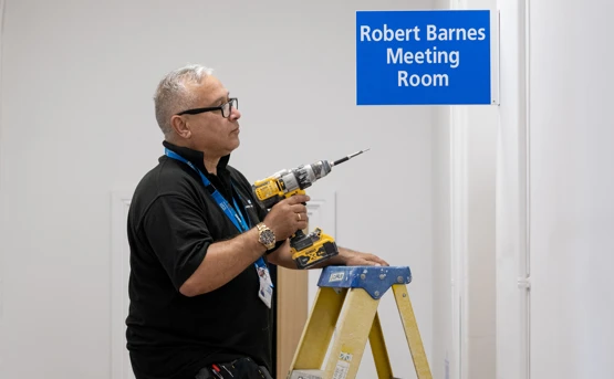 A man stands on a ladder using a power drill near a blue sign that reads Robert Barnes Meeting Room in a white-walled room.