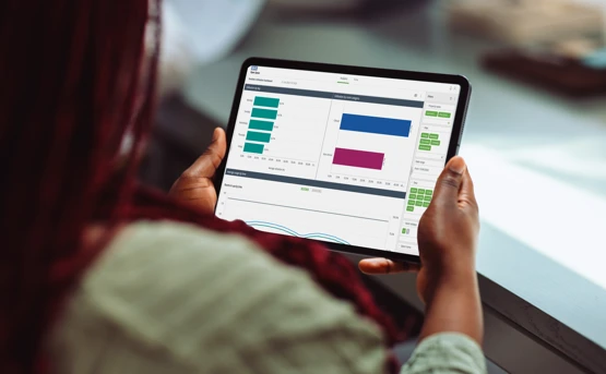 A person holds a tablet displaying data charts and graphs, including bar charts in green and blue, while sitting at a desk. The screen shows analytics or performance metrics in a dashboard interface.