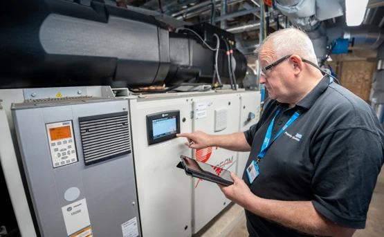 A man wearing glasses and a black polo shirt uses a touchscreen on industrial equipment while holding a tablet in a machinery-filled room.