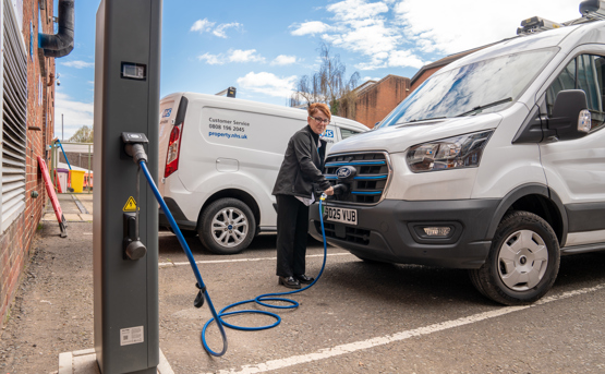 A person charges a white van at an electric vehicle charging station in a parking lot, with another white van and some buildings visible in the background.