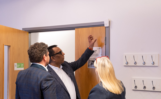 Three people stand by a wooden door; one man gestures toward a white sensor device above the door while the others observe. Several coat hooks are mounted on the wall nearby.