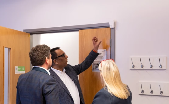 Three people stand by a wooden door; one man gestures toward a white sensor device above the door while the others observe. Several coat hooks are mounted on the wall nearby.