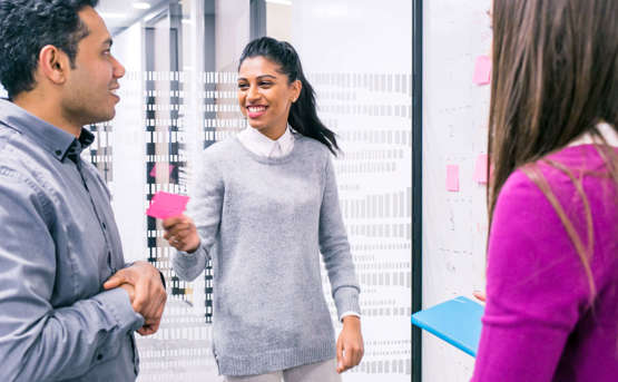 3 colleagues in an office environment working on a collaborative task. One of the colleagues is smiling at her colleague while's she's handing him a pink post-it