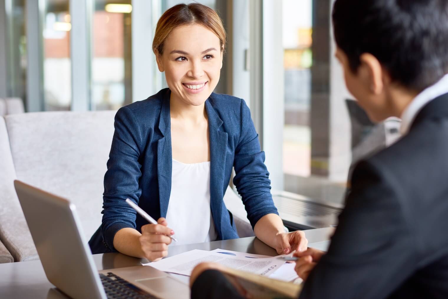 a smiling woman signs paper in front of a man wearing a suit.